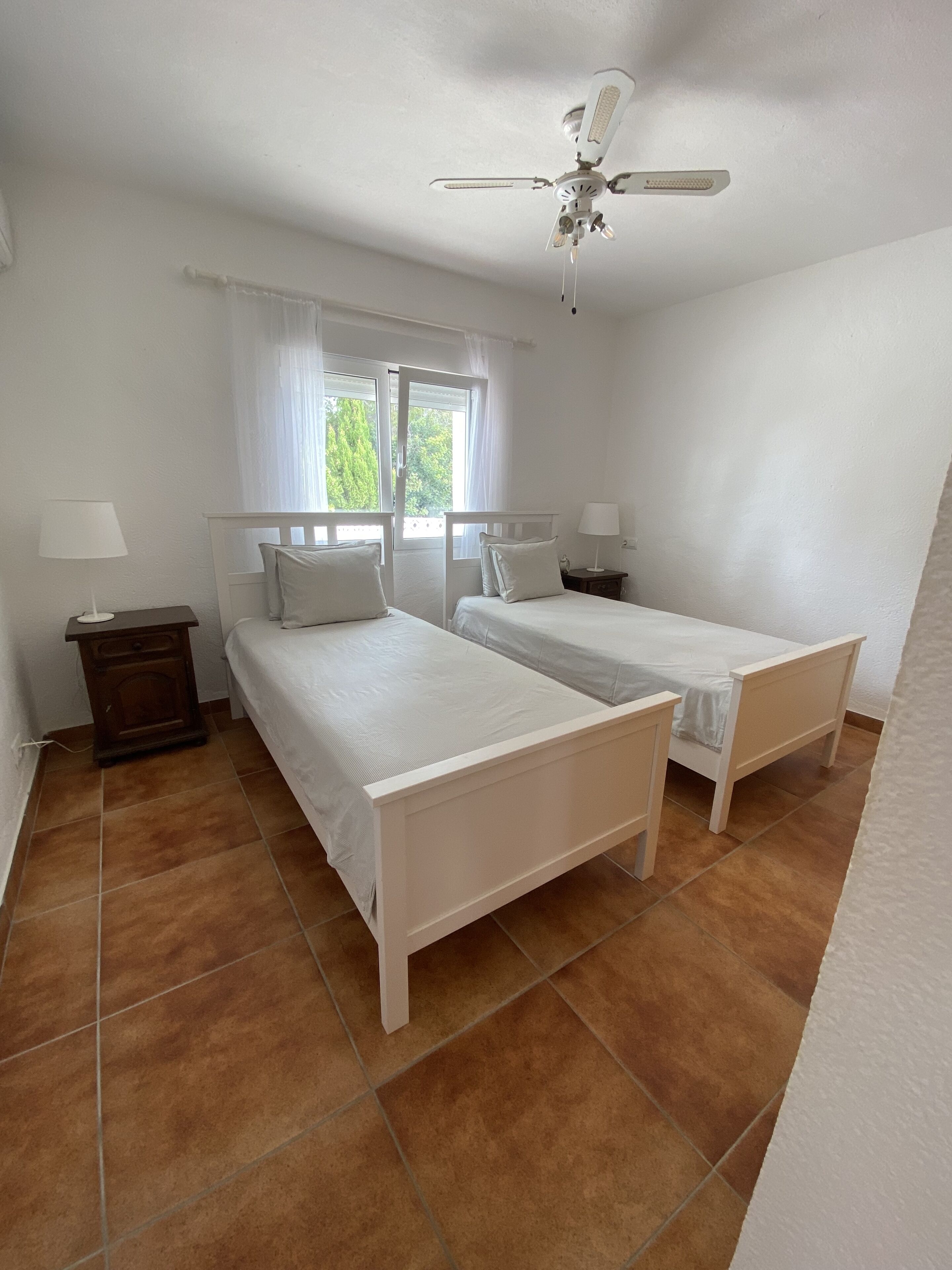 Een slaapkamer met twee enkele bedden, een plafondventilator, een raam met gordijnen en een nachtkastje met een lamp.