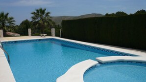 Una piscina rectangular con una sección curva, rodeada por un muro bajo y un seto.