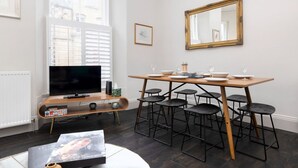 A modern living room with a TV, wooden dining table, black bar stools, and a large mirror on the wall.