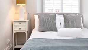 A neatly made bed with a grey blanket, a bedside table with a lamp, and a window with blinds.
