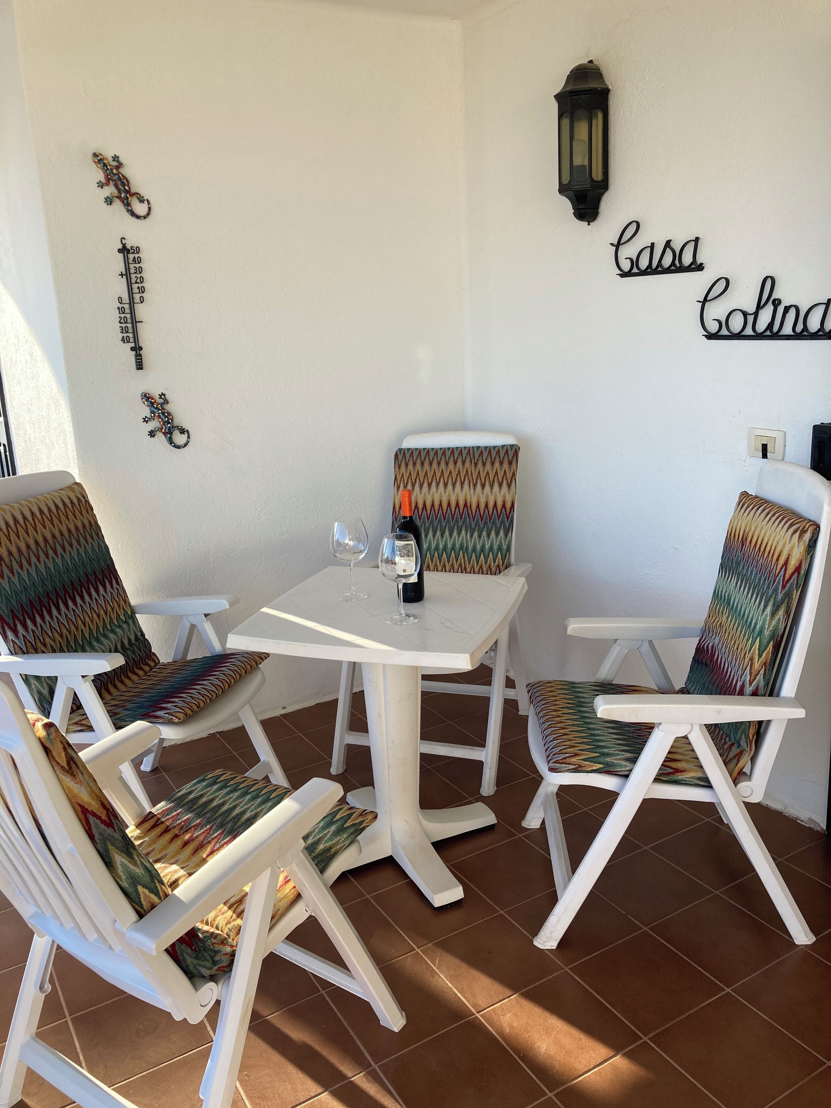 A small, cozy outdoor seating area with white chairs and a table, decorated with colorful cushions and a wall-mounted lamp.