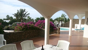 Un espace extérieur abrité, aménagé pour les repas, avec des chaises blanches et une table pour deux, offrant une vue sur une piscine et une végétation luxuriante.