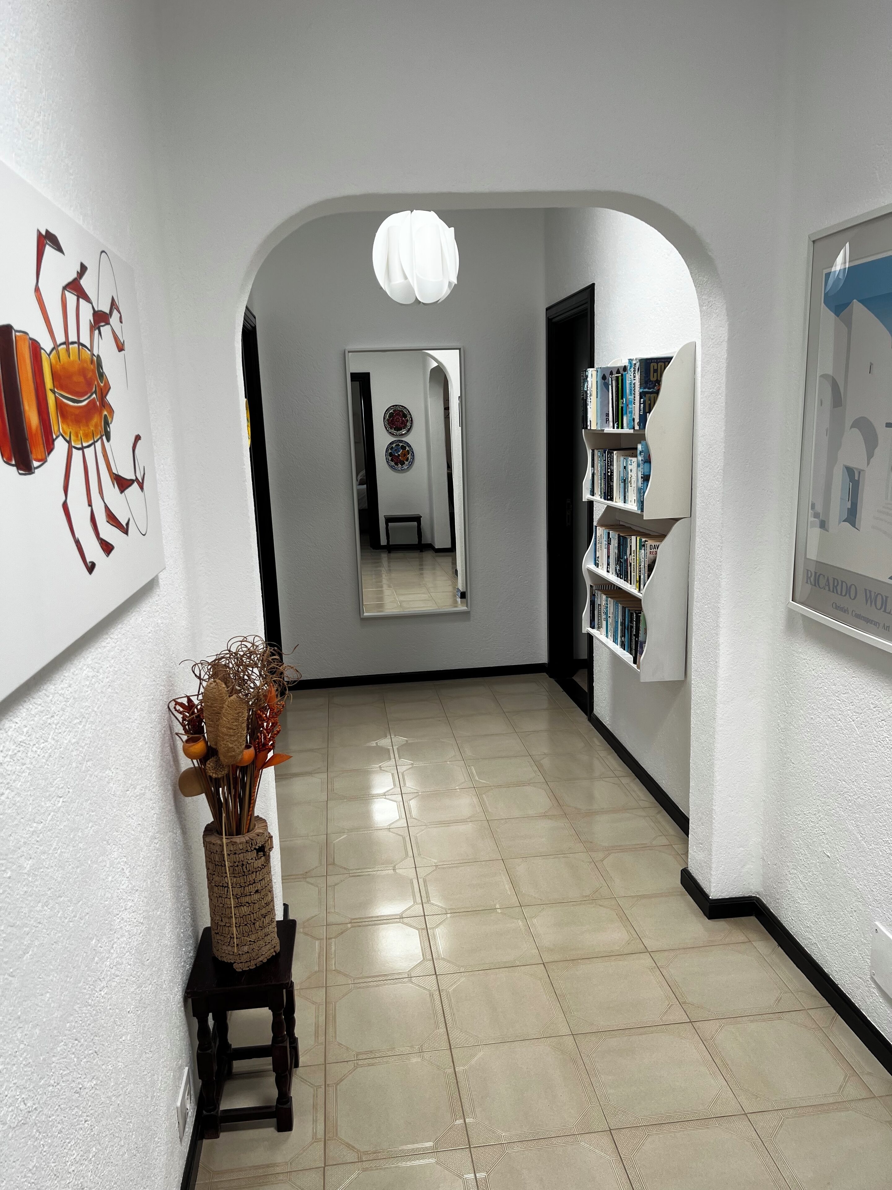A hallway with a tiled floor, a bookshelf, a decorative vase, and a framed picture on the wall.