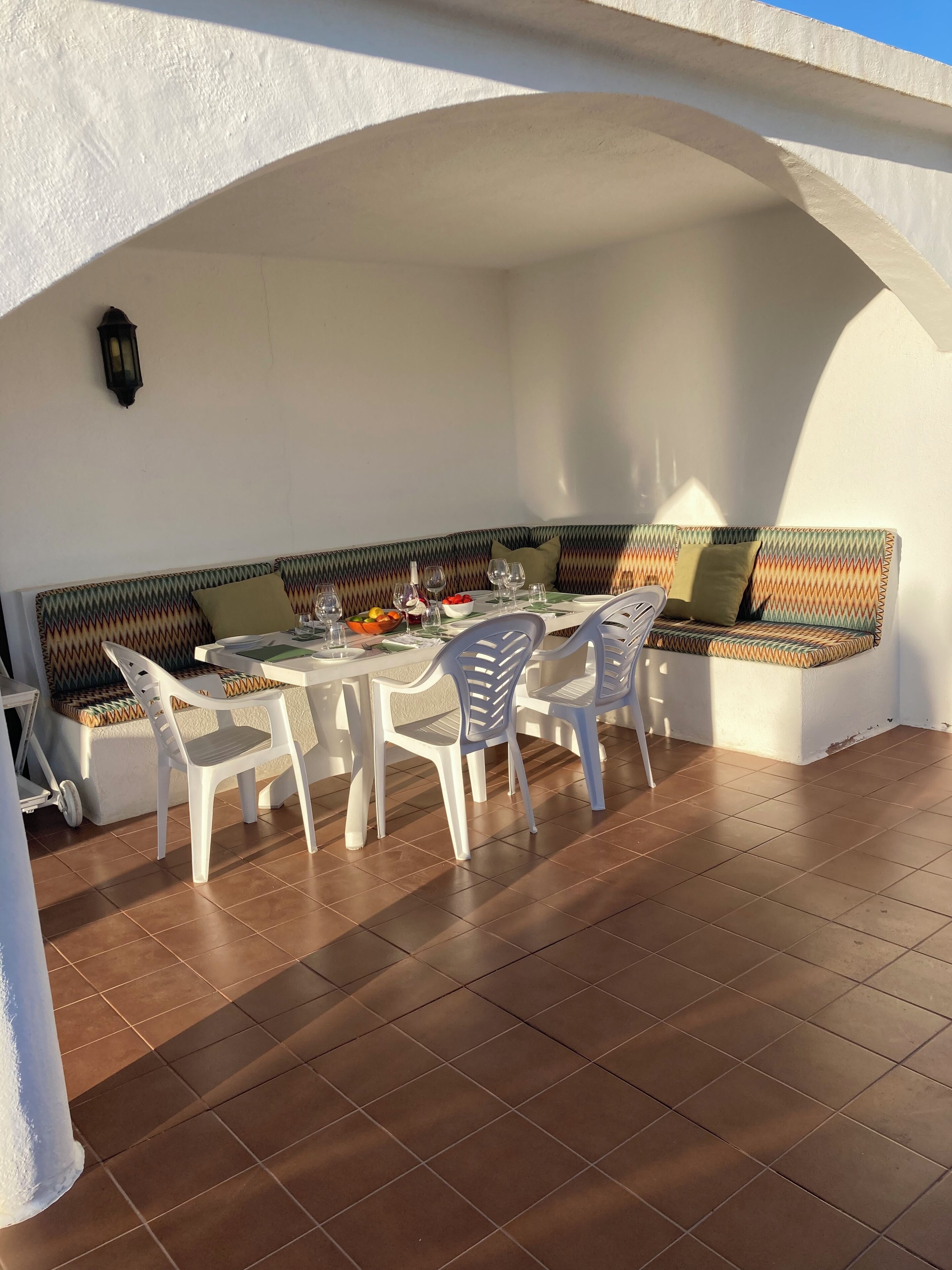 A covered outdoor dining area with a white table and chairs, a bench with patterned cushions, and a tiled floor.