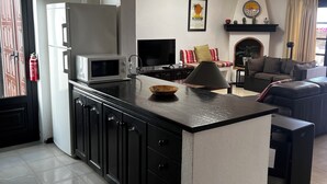 Une cuisine moderne avec un îlot central, des meubles de couleur noire et un four à micro-ondes. On y trouve également une cheminée surmontée d’une étagère, ainsi qu’un canapé dans l’espace salon.