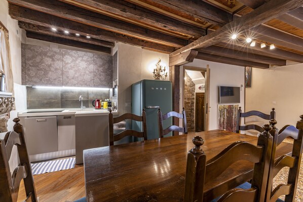 A dining area with a wooden table and chairs, a kitchen with a refrigerator, and a stone wall.