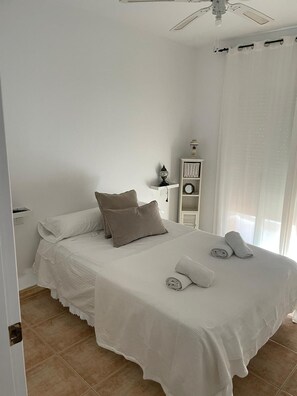 A neatly made bed with white linens and two pillows, a small bedside table with a lamp, and a white wardrobe in a bright room with a ceiling fan.
