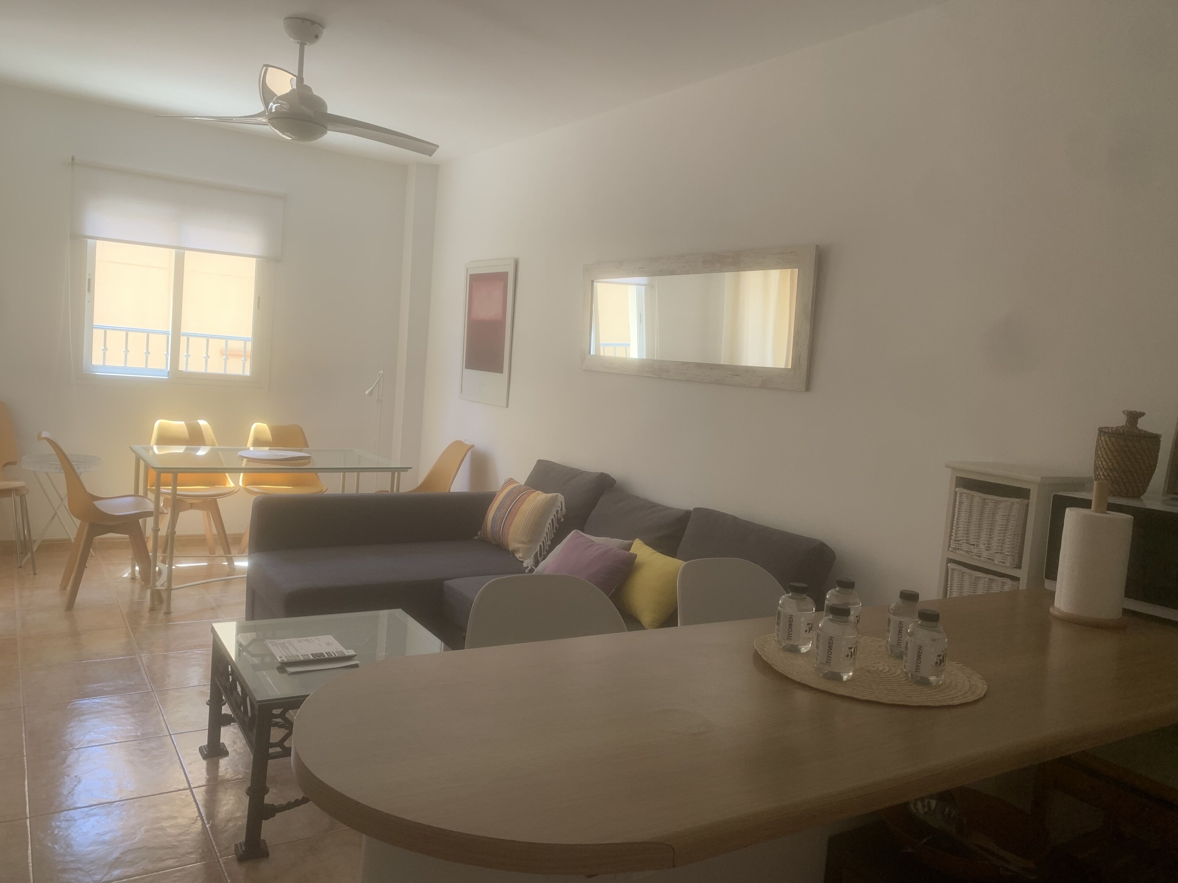 A living room with a dark-colored sofa, a glass coffee table, and a dining area with yellow chairs and a glass table.