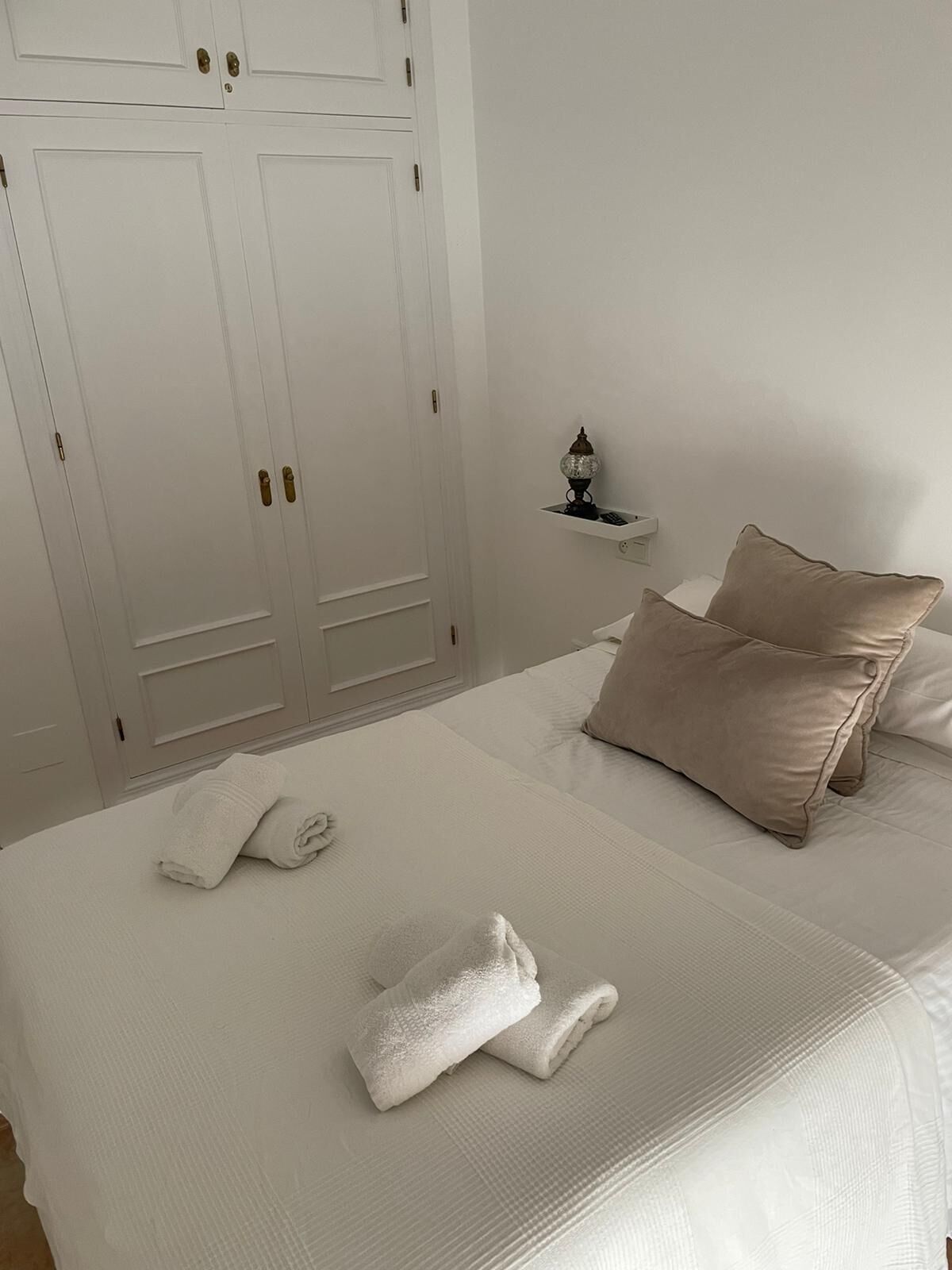 A neatly made bed with white linens and two rolled towels, a bedside table with a lamp, and a large wardrobe with multiple doors.
