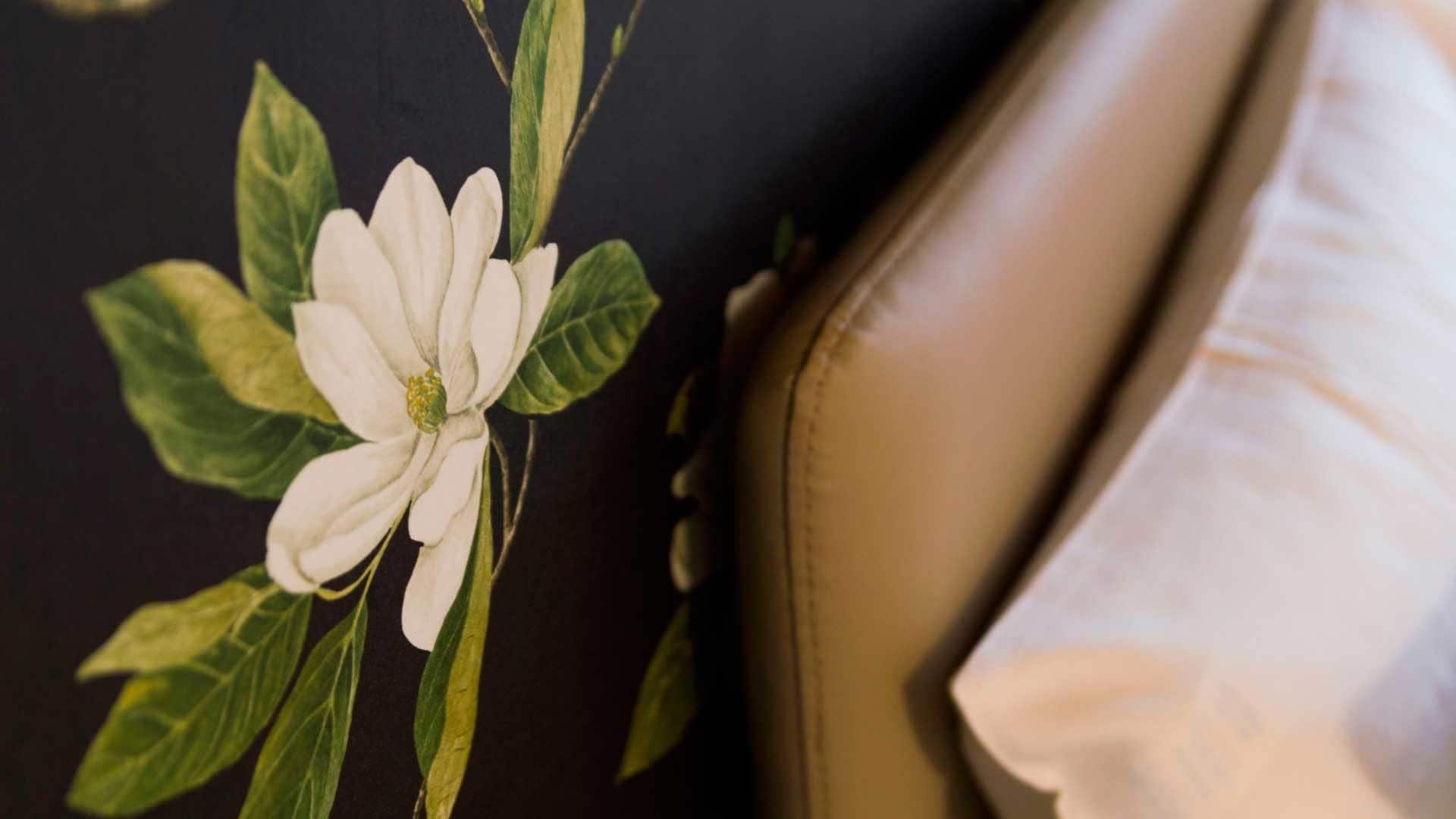 Tæt på en væg med blomstermønster, en brun lædersofa og en hvid pude.