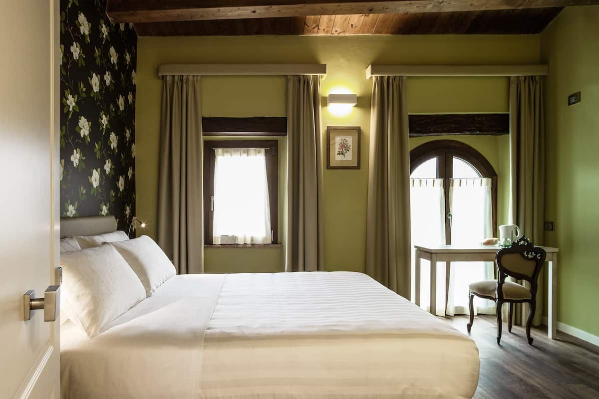 A bedroom with a large bed, two windows with curtains, a desk with a chair, and a framed picture on the wall.