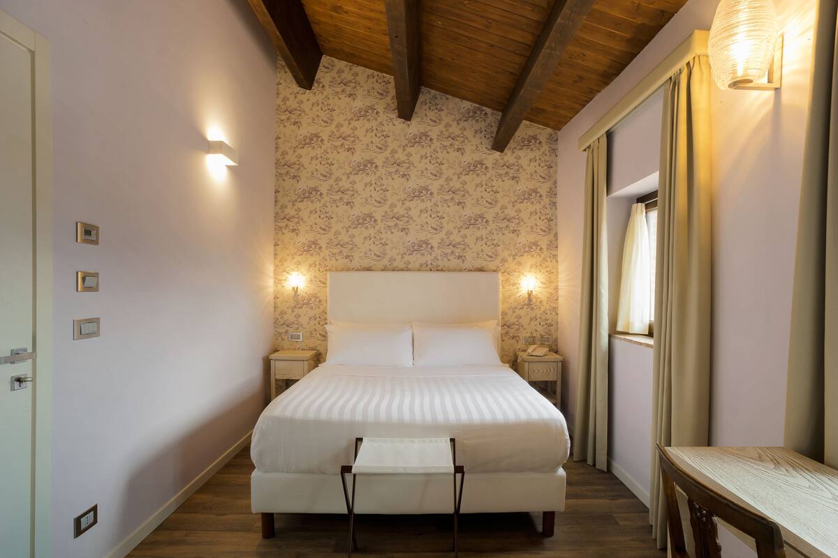 A bedroom with a bed, bedside tables, a nightstand, a wooden ceiling, and a window with curtains.