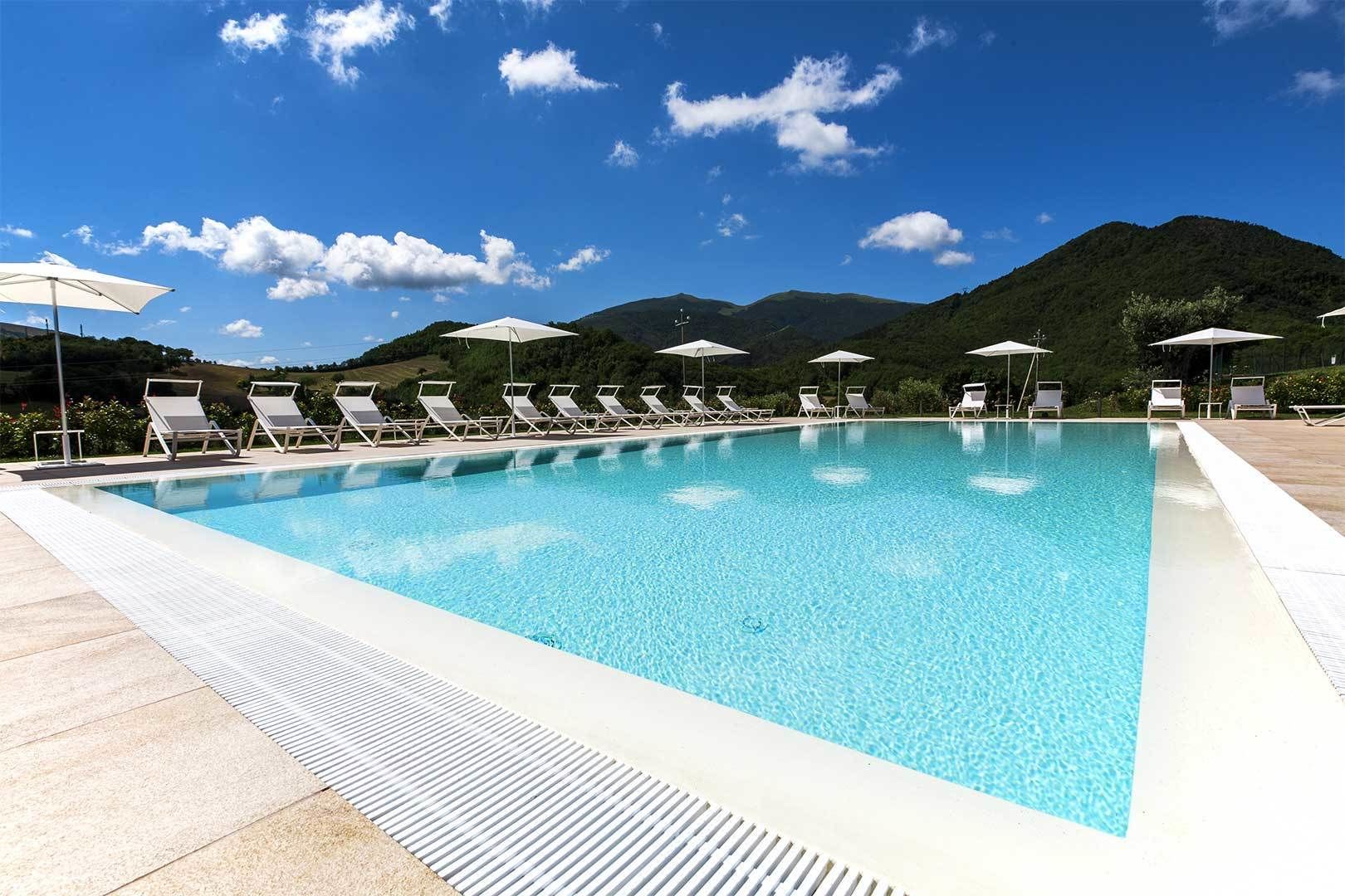 A rectangular swimming pool with clear blue water, surrounded by white lounge chairs and umbrellas, set against a backdrop of green hills and a clear blue sky.