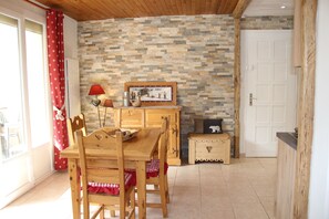 Een houten eettafel met stoelen, een rood-wit geruit tafelkleed, een rode lamp, een lijst met een afbeelding en een houten dressoir tegen een stenen muur.