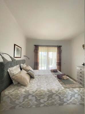 A bedroom with a bed, a dresser, a window with curtains, and a wall-mounted lamp.