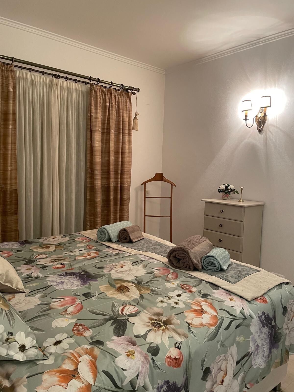 A bedroom with a floral bedspread, a bedside table with a lamp, and a wardrobe.