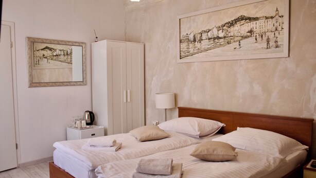 A double bed with white linens and pillows, a bedside table with a lamp, a wardrobe, and two framed artworks on the wall.