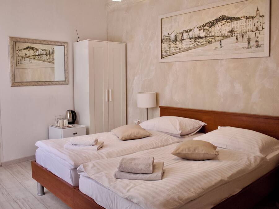 Un lit double avec du linge de lit blanc et des oreillers, une table de chevet avec une lampe, une armoire et deux œuvres d’art encadrées accrochées au mur.