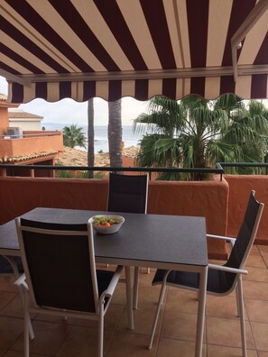 Une terrasse avec une table et des chaises, un saladier rempli de fruits et une vue sur la plage et les palmiers.