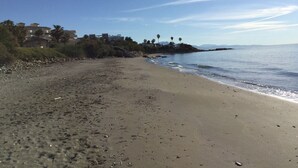 A sandy beach with a clear sky, a rocky shoreline, and buildings in the background.