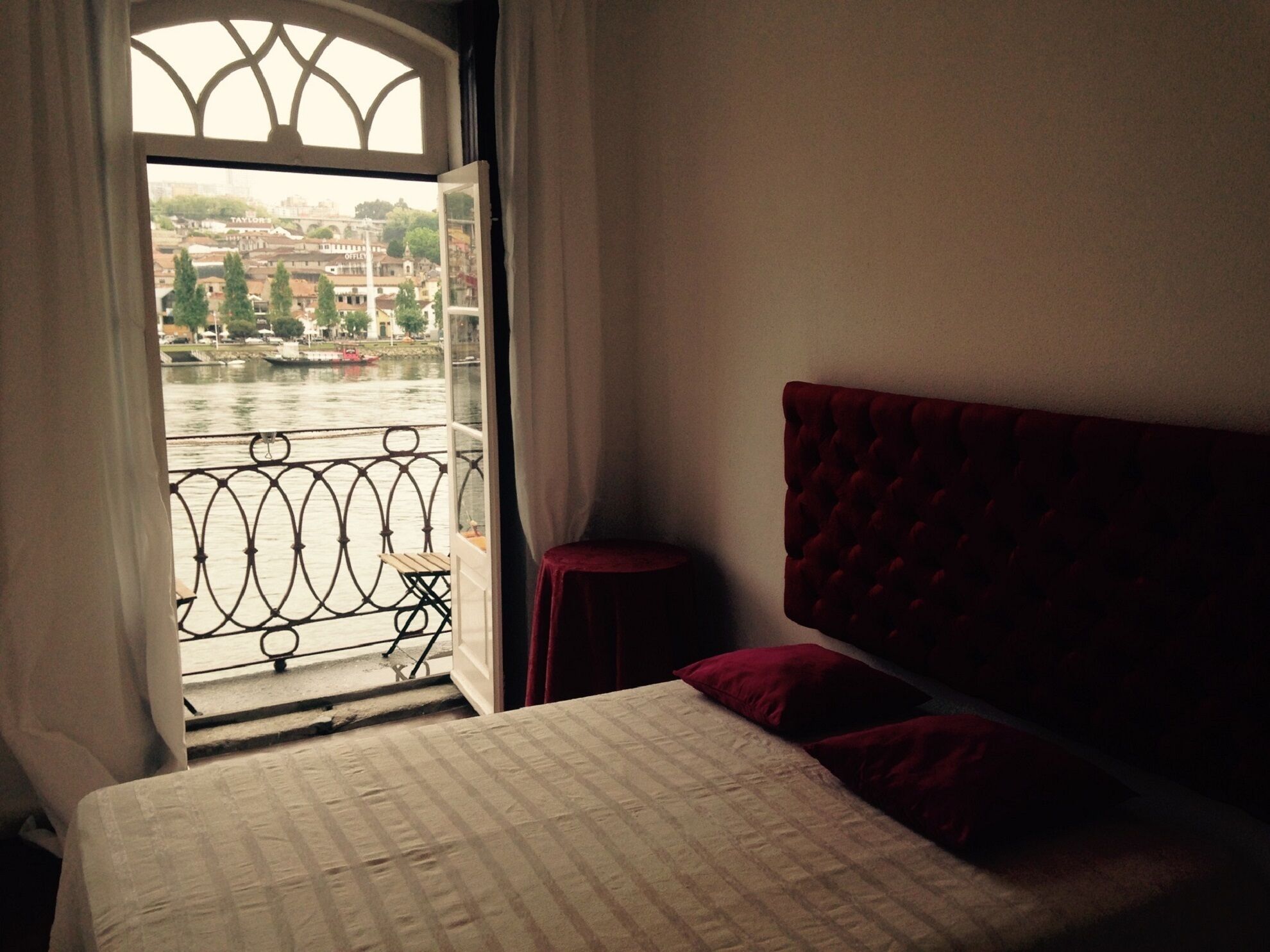 A bedroom with a bed, a red headboard, a small table, and a view of a river and buildings through a window.