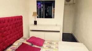 Un dormitorio con un cabecero rojo con capitoné, una cama con colcha de colores y diseños, dos almohadas rosas, un tocador blanco con espejo y una mesita de noche con lámpara.