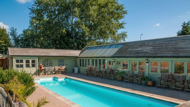 A pool area with a clear blue pool, wooden deck chairs, and a building with a green facade and large windows. - Beautiful cabin near Bath with heated swimming pool sleeps up to 17 people (Bristol)