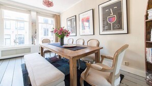 Un coin repas avec une table en bois, quatre chaises, un banc et un vase de fleurs. Des tableaux encadrés ornent le mur, et une bibliothèque se trouve à droite.
