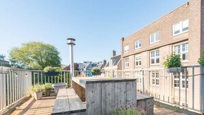 Une terrasse en bois agrémentée de jardinières et d’un banc, entourée d’une clôture en métal et de bâtiments résidentiels.