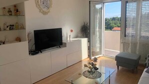 Een moderne woonkamer met een flatscreen televisie, een glazen salontafel met decoratieve objecten, een grijze bank en een balkon met uitzicht op groen.