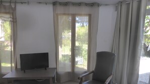 Una habitación con televisión sobre un mueble bajo, una silla y un amplio ventanal con cortinas.