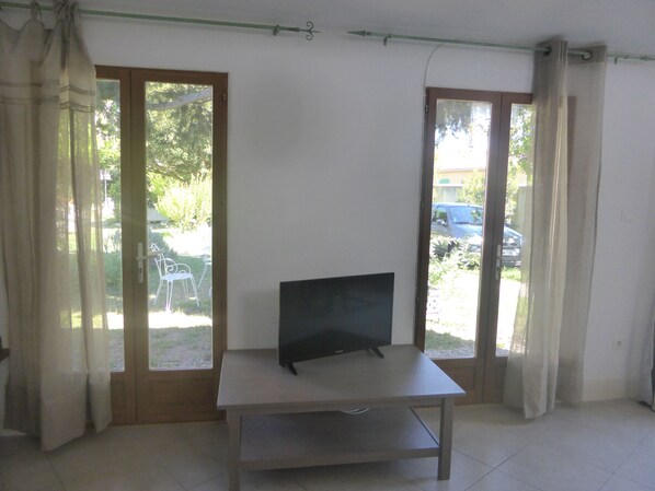 A room with a TV on a table, large windows with curtains, and a view of the outside. - Studio in Mas provençal in AVIGNON (2 people) (Avignon)