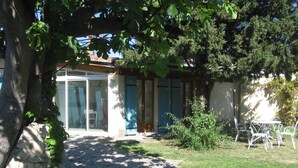 Una casa con puerta de cristal, un pequeño patio con mesa y sillas, y un jardín con plantas y un árbol.