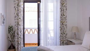 A bedroom with a bed, a nightstand, a lamp, a vase with a plant, and a view of the outside through a door.