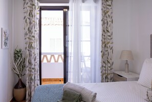 A bedroom with a bed, a nightstand, a lamp, a vase with a plant, and a view of the outside through a door.