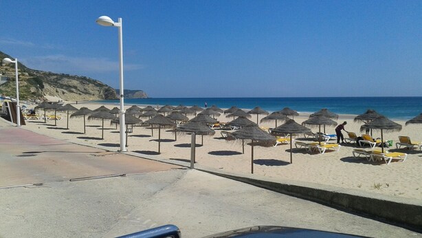 A beach with rows of sun loungers and thatched umbrellas, a clear sky, and a calm sea.