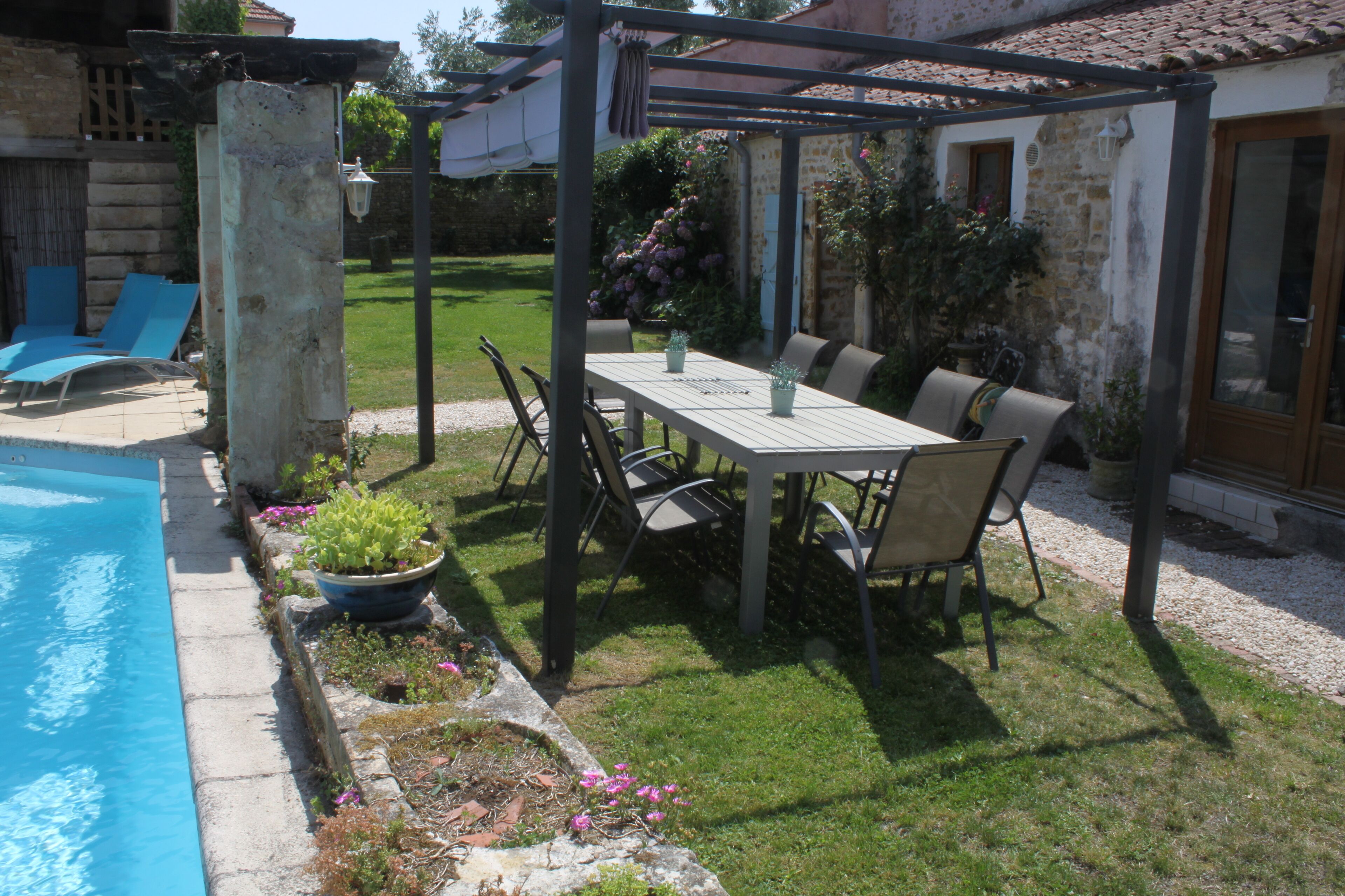 A covered outdoor dining area with a table and chairs, a pool, and a garden with flowers and plants.