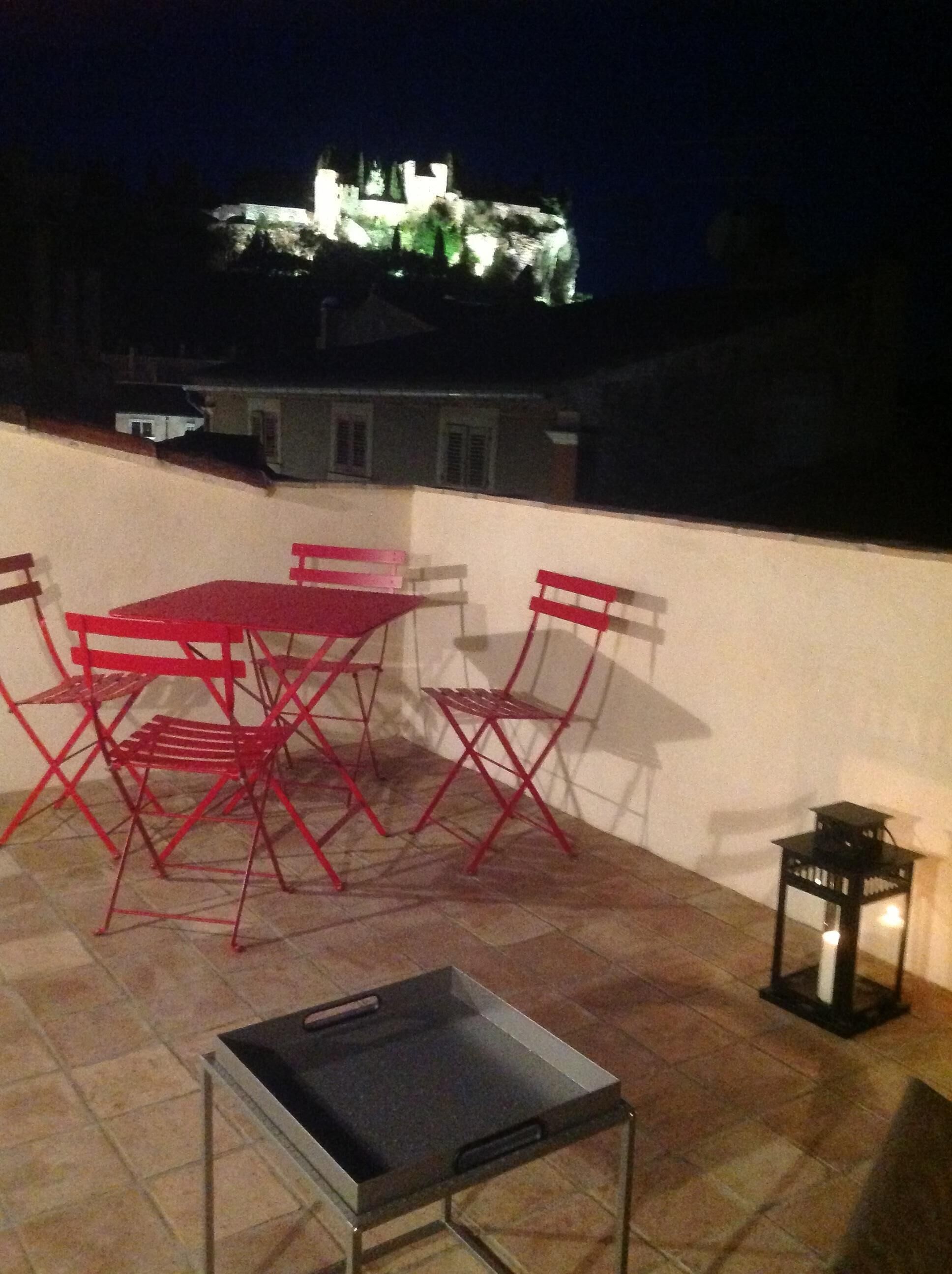 A rooftop terrace with a table and chairs, a lantern, and a view of a castle at night.