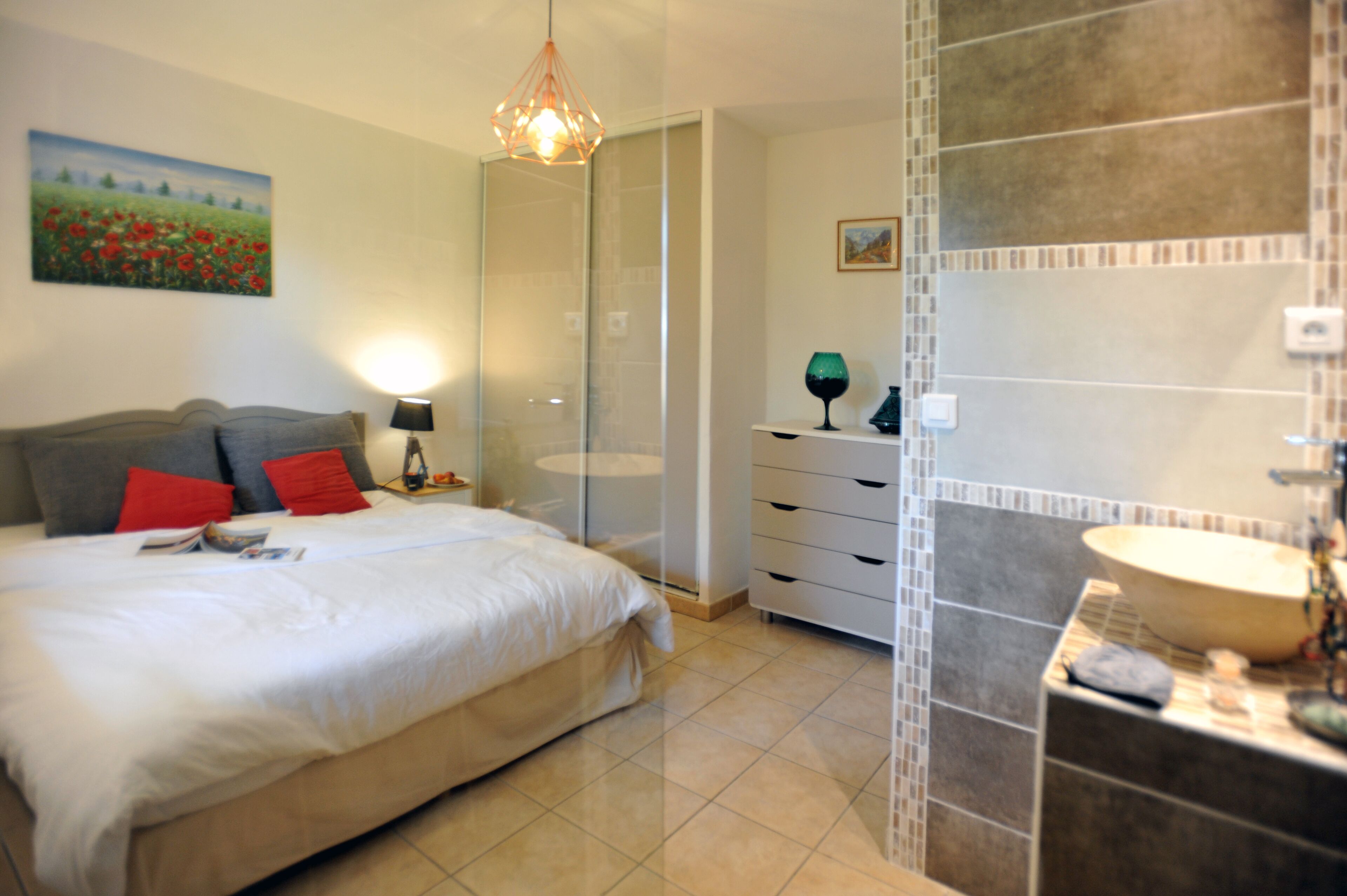 A bedroom with a bed, a nightstand, a lamp, a painting, a mirror, and a bathroom with a sink and tiles.