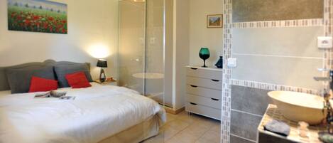 A bedroom with a bed, a nightstand, a lamp, a painting, a mirror, and a bathroom with a sink and tiles.