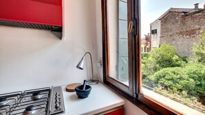 A kitchen with a gas stove, a window with a view of a garden and buildings, and a red cabinet. - BRAGORA SMOOCHY ATTIC VENICE M0270423479 (Venice)