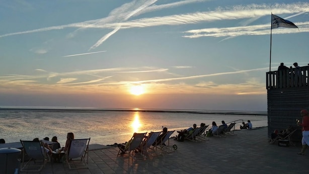 Sonnenuntergangsblick von einer Terrassenanlage am Strand mit Liegestühlen und einer Fahnenstange.