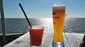 Dos vasos de cerveza sobre una mesa de madera con vista al mar y una cometa a lo lejos.