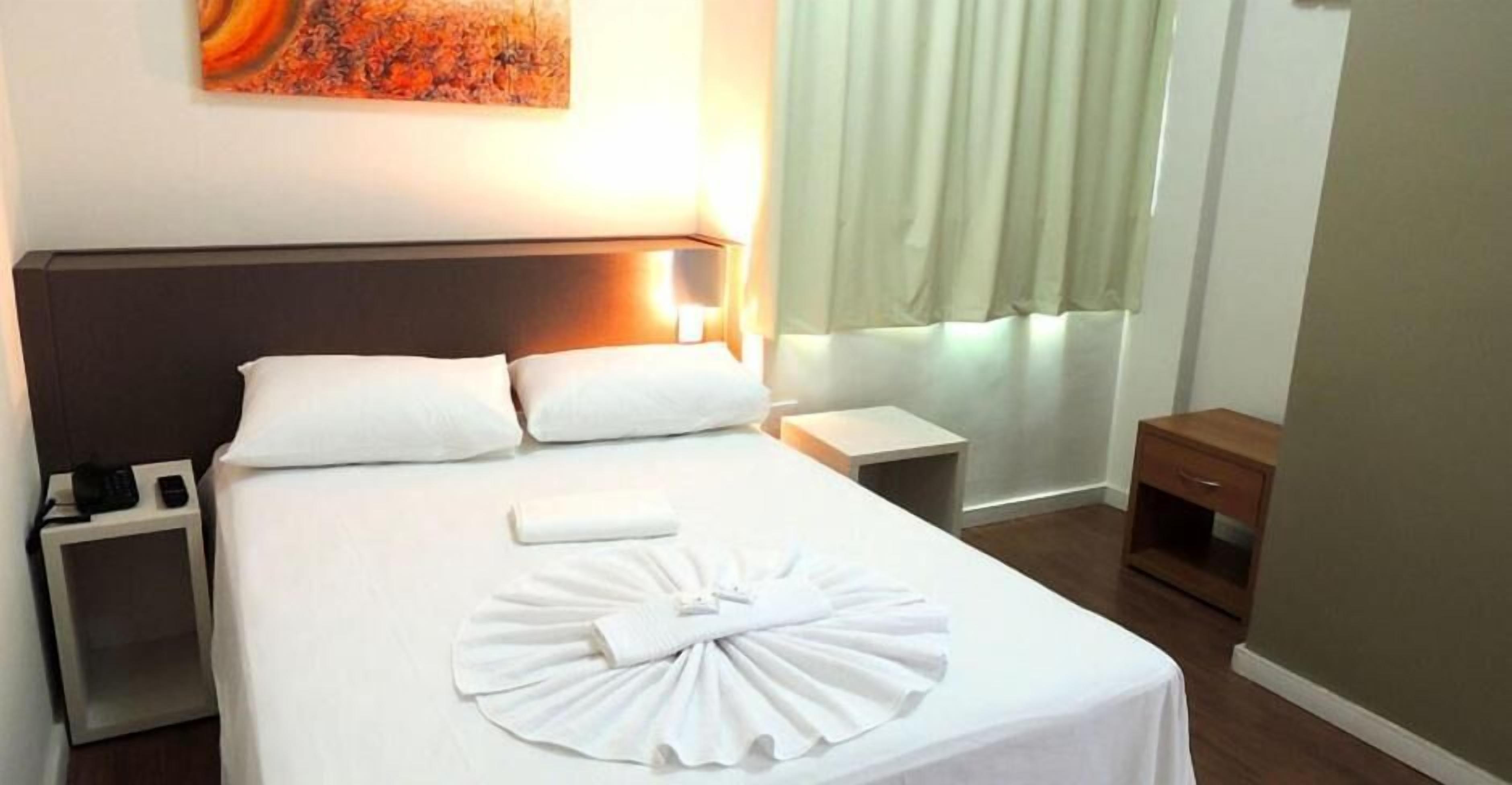 Una cama bien hecha con sábanas blancas, una mesita de noche con lámpara y una pequeña cómoda de madera.