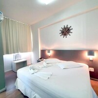 Cama bem arrumada com lençóis e toalhas brancas, mesa de cabeceira com abajur e um objeto decorativo na parede.