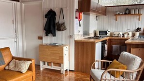 A cozy room with a wooden ceiling, a white door, a brown sofa, a wicker chair, a woven ottoman, and a kitchen area with a microwave and toaster. - SLOY CABIN is a charming cabin in a stunning loch side location, with hot tub. (Taynuilt)