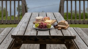 Una mesa de picnic de madera con una tabla de quesos, uvas y pan, con vistas a un lago y colinas al fondo.