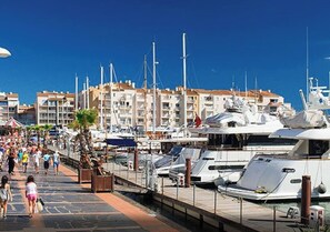 A marina with several yachts docked, a pedestrian walkway, and a building with balconies in the background.