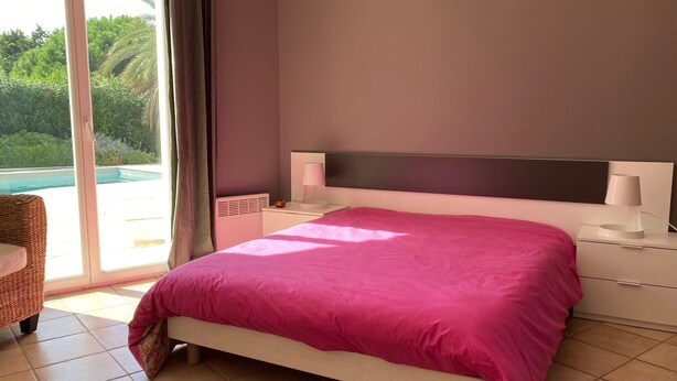 A bedroom with a large bed, a nightstand, a lamp, and a view of a swimming pool through a glass door.