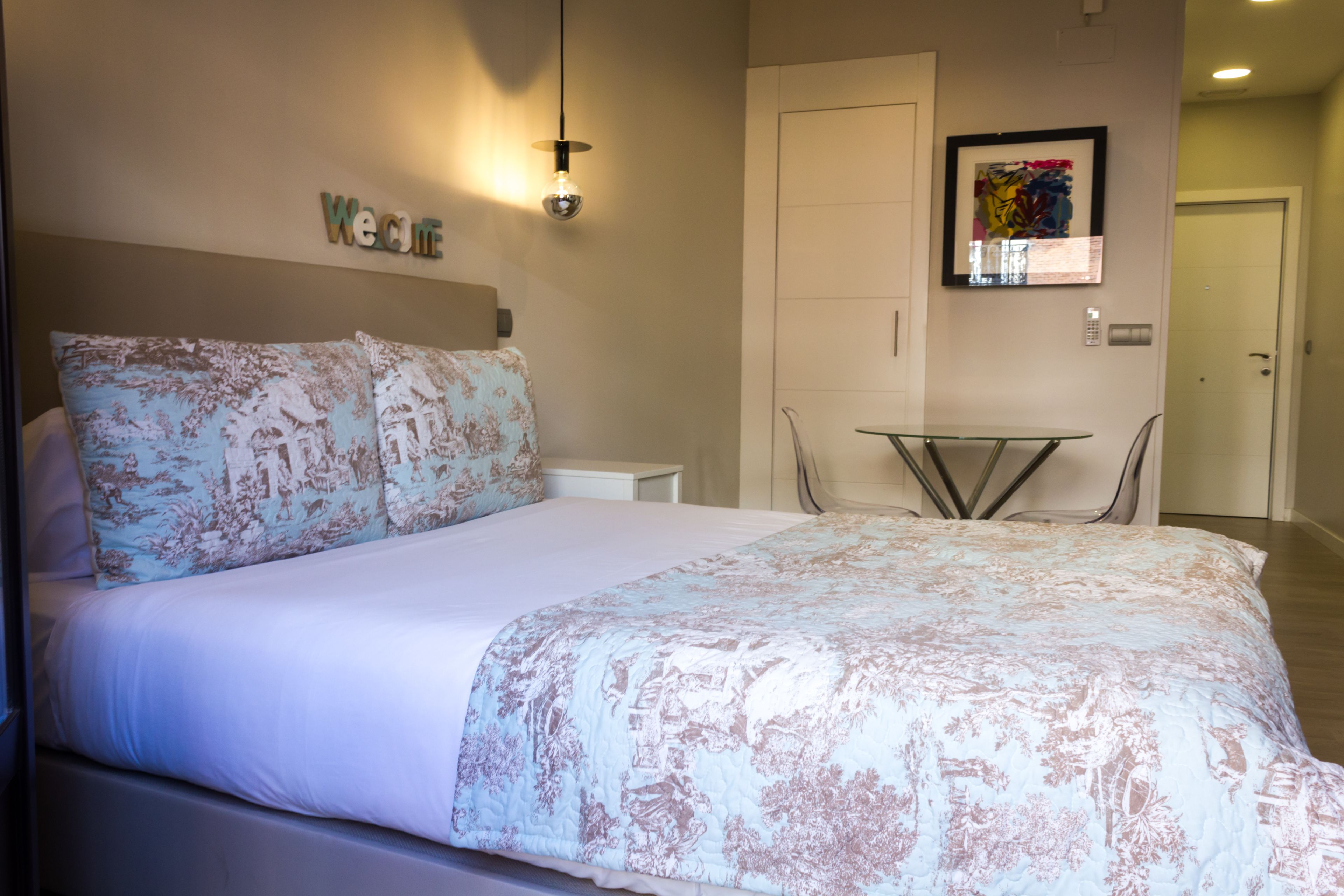 A neatly made bed with floral bedding, a small table, and a framed artwork on the wall.